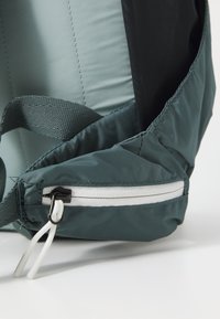 Grön nylonväska med en texturerad yta, prydd med en vit dragkedjedetalj och justerbar rem. Kompakt och lätt design med mjuka kanter.