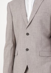 Blazer cinza claro, texturizado, com fecho de dois botões, lapelas recortadas e um bolso no peito. Projetado para um ajuste sob medida.