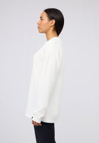 Weißes, strukturiertes Langarmshirt mit Rundhalsausschnitt und lockerer Passform, ausgestattet mit engen Manschetten und einem seitlichen Etikett.
