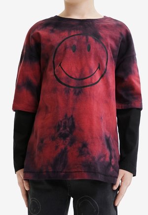 Kind draagt een rood-zwart tie-dye T-shirt met korte mouwen met een groot zwart lachend gezicht, gelaagd over een zwart longsleeve T-shirt en een zwarte broek.