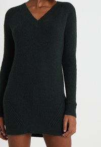 Vestido de suéter de punto verde oscuro con escote en V, mangas largas, textura acanalada y detalles laterales. El dobladillo llega por encima de los muslos.