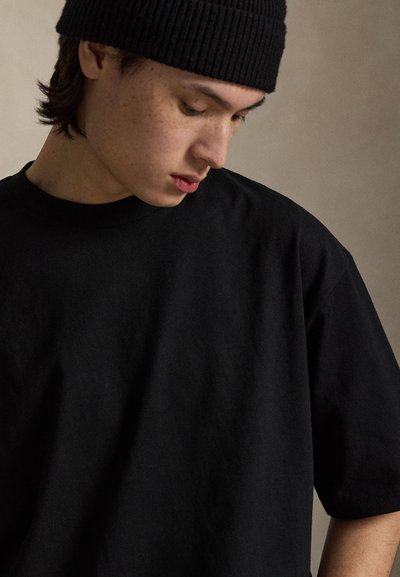 Νεαρό άτομο με φακίδες, φοράει μαύρο σκουφάκι και μαύρο μπλουζάκι, κοιτάζει προς τα κάτω μπροστά σε απλό φόντο.