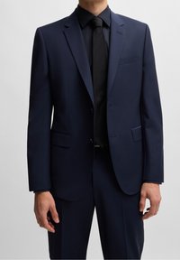 Abito sartoriale blu navy con giacca monopetto, rever affilati e due tasche frontali, abbinato a una cravatta nera e una camicia nera.