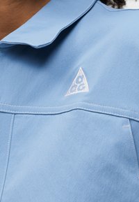Κοντινή λήψη του γιακά και της περιοχής του στήθους ενός ανοιχτό μπλε ενδύματος με λευκό τριγωνικό λογότυπο κεντημένο στο ύφασμα.