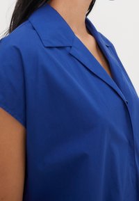 Donna che indossa una camicia a maniche corte di colore blu reale con colletto e chiusura a bottoni su uno sfondo semplice.