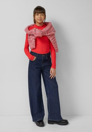 Czerwony top z długim rękawem w połączeniu z szerokimi jeansami z ciemnego denimu. Na ramionach zawinięty czerwono-biały sweter w kratę. Czarne buty.