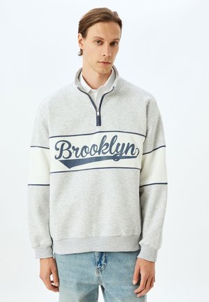 Lys grå sweatshirts med halv-zip krave, marineblå "Brooklyn" tekst, cremefarvede accenter og ribbede manchetter. Blødt, tekstureret stof.