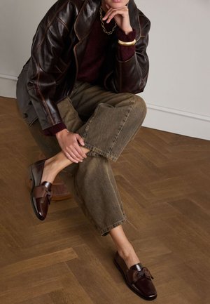 Mocassins en cuir brun avec un accent en nœud, présentant une finition brillante. Portés avec un pantalon large olive, à revers, sur un sol en bois.