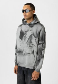 Grijze tie-dye hoodie met een voorzak, voorzien van zwarte grafische tekst. Model draagt donkere jeans en rechthoekige zonnebril. Glad textuur.
