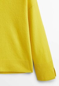Manche et ourlet d'un pull en tricot jaune avec poignets et bord inférieur côtelés sur fond blanc.