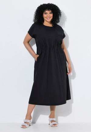 Frau mit lockigem Haar, die ein schwarzes Midikleid mit Taschen und weiße Sandalen mit hohen Absätzen trägt, steht vor einem weißen Hintergrund.
