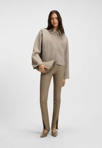Cappotto beige corto con colletto a punta, abbinato a pantaloni marroni aderenti con spacchi laterali. Il modello tiene una elegante borsa color nocciola.