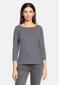 Gestreiftes Langarmoberteil in Navy und Weiß mit einem Bootsausschnitt, aus weichem Material gefertigt. Kombiniert mit grauen Jeans zeigt das Modell einen lässigen Schnitt.