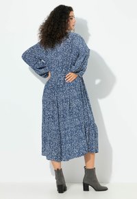 Kobieta o kręconych włosach, ubrana w niebiesko-białą, wzorzystą sukienkę midi z długim rękawem oraz szare botki, stojąca z rękami na biodrach.