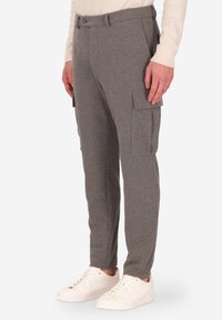 Uomo che indossa pantaloni cargo slim-fit grigi con tasche laterali e sneakers bianche, in piedi di fronte a uno sfondo bianco uniforme.