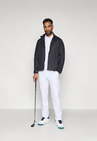 Svart lättviktsjacka, vit pikétröja och vita byxor. Modellen håller i en golfklubba och bär vita skor med blå accenter. Enkel design och passform.