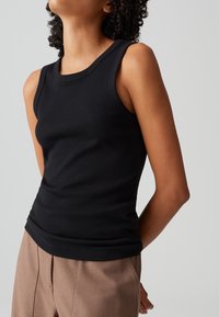 Schwarzes geripptes Tanktop mit ärmellosem Design und Rundhalsausschnitt. Kombiniert mit hellbraunen Hosen, die eine figurbetonte Silhouette zeigen.