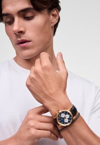 Orologio cronografo dorato con quadrante blu, cinturino in pelle nera e accentuazioni argentate. Indossato su un polso maschile su uno sfondo bianco semplice.