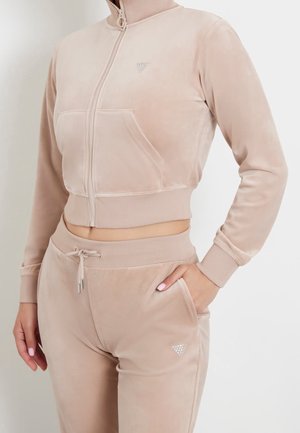 Kobieta ubrana w beżową, aksamitną, zapinaną na zamek krótka kurtkę i pasujące spodnie ze ściągaczem, z ręką w kieszeni, na białym tle.