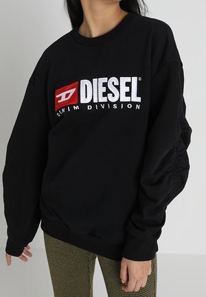 Person iført en sort Diesel Denim Division sweatshirt med stort hvidt og rødt logo, kombineret med grønne ternede bukser, står indendørs.