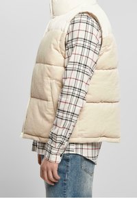 Beige kordfløyels puffer-vest med en hvit polstret krage, med glidelås og sidelommer, båret over en svart og hvit rutet skjorte.