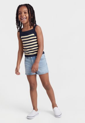 Χαμογελαστή κοπέλα με πλεξούδες, φορώντας μπλε ναυτικό και μπεζ ριγέ αμάνικο τοπ, τζιν σορτς και λευκά παπούτσια τύπου slip-on, στέκεται μπροστά σε απλό φόντο.