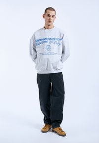 Gray sweatshirt with blue text and graphics, relaxed fit, paired with black pants and tan sneakers. Text includes brand name and slogan details.