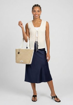 Femme souriante aux cheveux tressés porte un gilet en maille sans manches couleur crème, un top blanc, une jupe midi bleu marine, des sandales noires à talons et tient un sac cabas tressé.