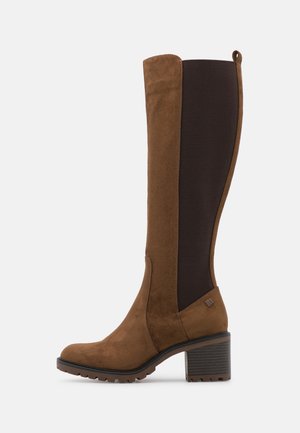 Stivale marrone in suede fino al ginocchio con pannello elastico laterale nero, suola carrarmato e tacco a blocco su sfondo bianco, vista di profilo.