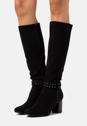 Bottes noires en daim jusqu'aux genoux avec un bout carré, un talon bloc haut et des accents cloutés autour de la cheville. Dotées d'une fermeture éclair latérale pour faciliter l'enfilage.