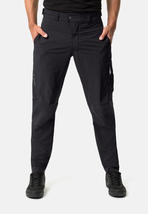 Pantaloni neri con una texture liscia, tasche laterali, cuciture doppie e vestibilità dritta, abbinati a sneakers nere per contrasto.