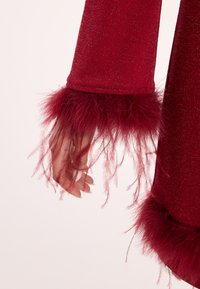 Rotes Kleid mit glitzernder Textur an den Ärmeln, die an den Bündchen mit flauschigen Federakzenten verziert sind, und eine Hand, die sanft gegen den Stoff ruht.