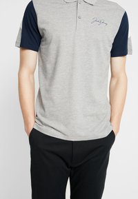 Šedé polokošile s námořnickými rukávy, dva knoflíky u límce a vyšitým logem. Měkká bavlněná směs s volným střihem a bočními rozparky.