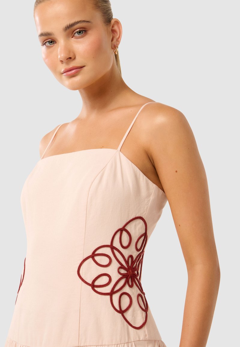 Lichtroze mouwloos jurk met ingewikkelde bordeauxrode bloemenborduurwerk aan de zijkanten, voorzien van een vierkante halslijn en een vloeiende rok.