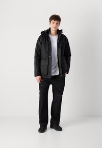 Fekete puffernadrág kapucnival, elülső zipzárral, két elülső zsebbel és matt felülettel, egy fehér ing és fekete nadrág fölött viselve.