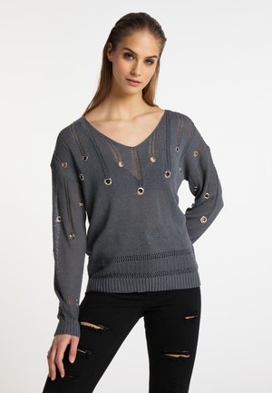 Jonge vrouw met lang haar draagt een grijze gebreide trui met metalen oogjes en zwarte gescheurde skinny jeans, staand tegen een witte achtergrond.