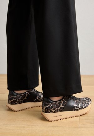 Personne portant un pantalon noir à jambes larges et des baskets compensées à imprimé léopard avec des semelles épaisses beige, debout sur un sol en bois.