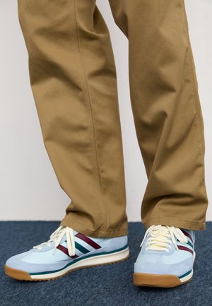 Personne portant des baskets bleu clair avec des rayures bordeaux et vertes et des lacets beiges, associées à un pantalon marron ample sur un tapis bleu.