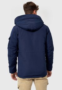 Veste isolante bleu marine avec une grande capuche, conçue avec une texture lisse et des poches avant, idéale pour le temps froid.