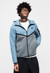 Ζακέτα Nike με φερμουάρ, με ανοιχτό μπλε πάνω μέρος και γκρι κάτω πάνελ, με μαύρες λεπτομέρειε ς, κουκούλα και τσέπη με φερμουάρ στον βραχίονα.