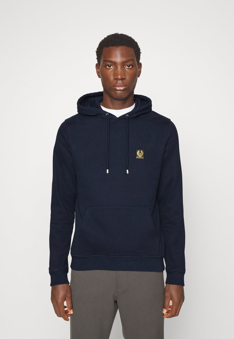 Σκούρο μπλε φούτερ από μαλακό ύφασμα, με καγκουρώ τσέπη, ρυθμιζόμενους κορδόνια και μια μικρή χρυσή λογοτύπωση στο στήθος.