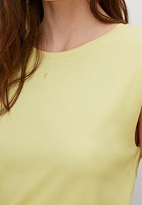 Žlutý žebrovaný tílko s kulatým výstřihem a bezramínkovým designem. Obsahuje jemně embosované logo poblíž límce. Hladký povrch.
