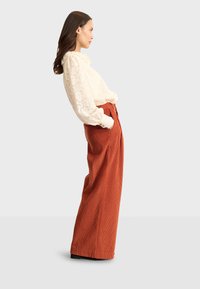 Blusa bianca in pizzo con collo a volant, abbinata a pantaloni in velluto a coste ruggine a vita alta e gamba larga. Visualizzazione di profilo.