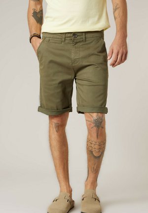 Homme portant un short olive retroussé, des chaussures beige à enfiler, des tatouages visibles sur les jambes et les bras, debout avec une main dans la poche.