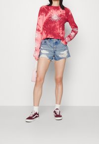 Červené a růžové batikované tričko s dlouhým rukávem se slunečním motivem, opotřebované džínové šortky, bílé ponožky, vínové tenisky a malá růžová kabelka.