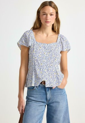 Blusa floreale con scollo quadrato, maniche a sbuffo corte e sfondo azzurro chiaro. Presenta motivi gialli e blu. Abbinata a jeans a vita alta.