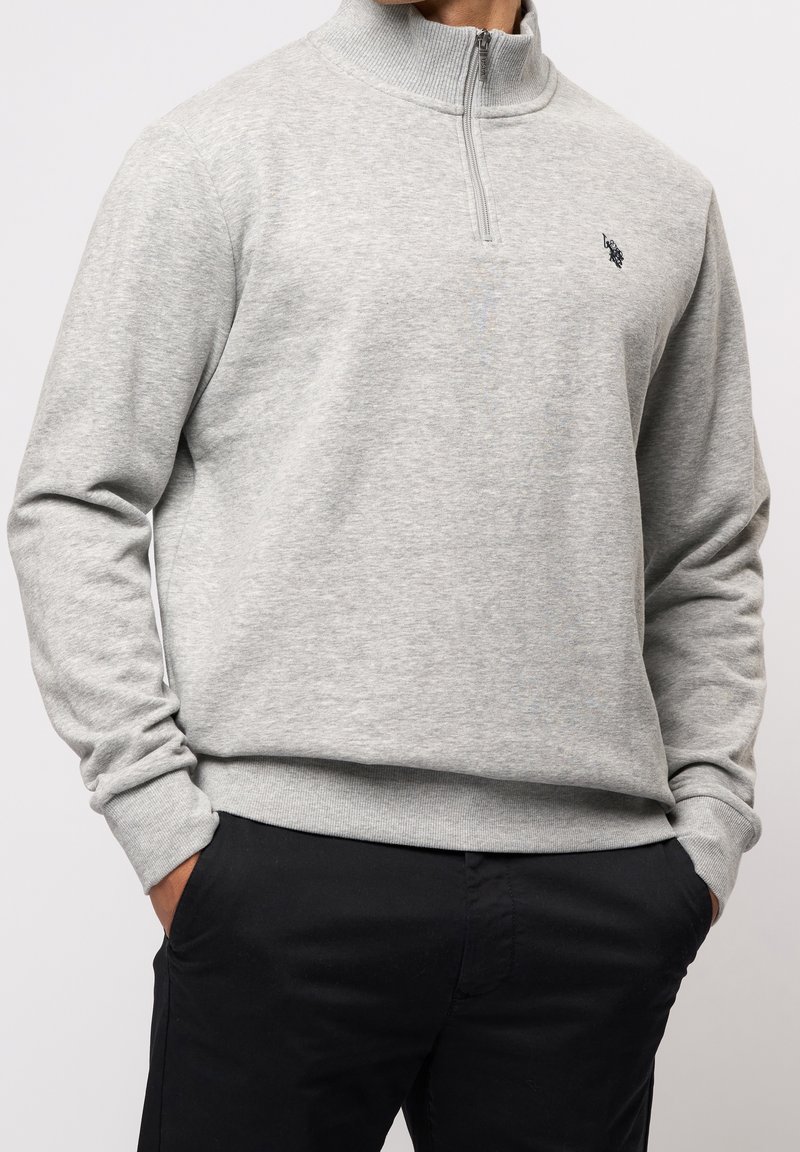 Homme portant un pull gris clair avec une fermeture éclair quart de hauteur et un petit logo sur la poitrine, ainsi qu'un pantalon noir, mains dans les poches.