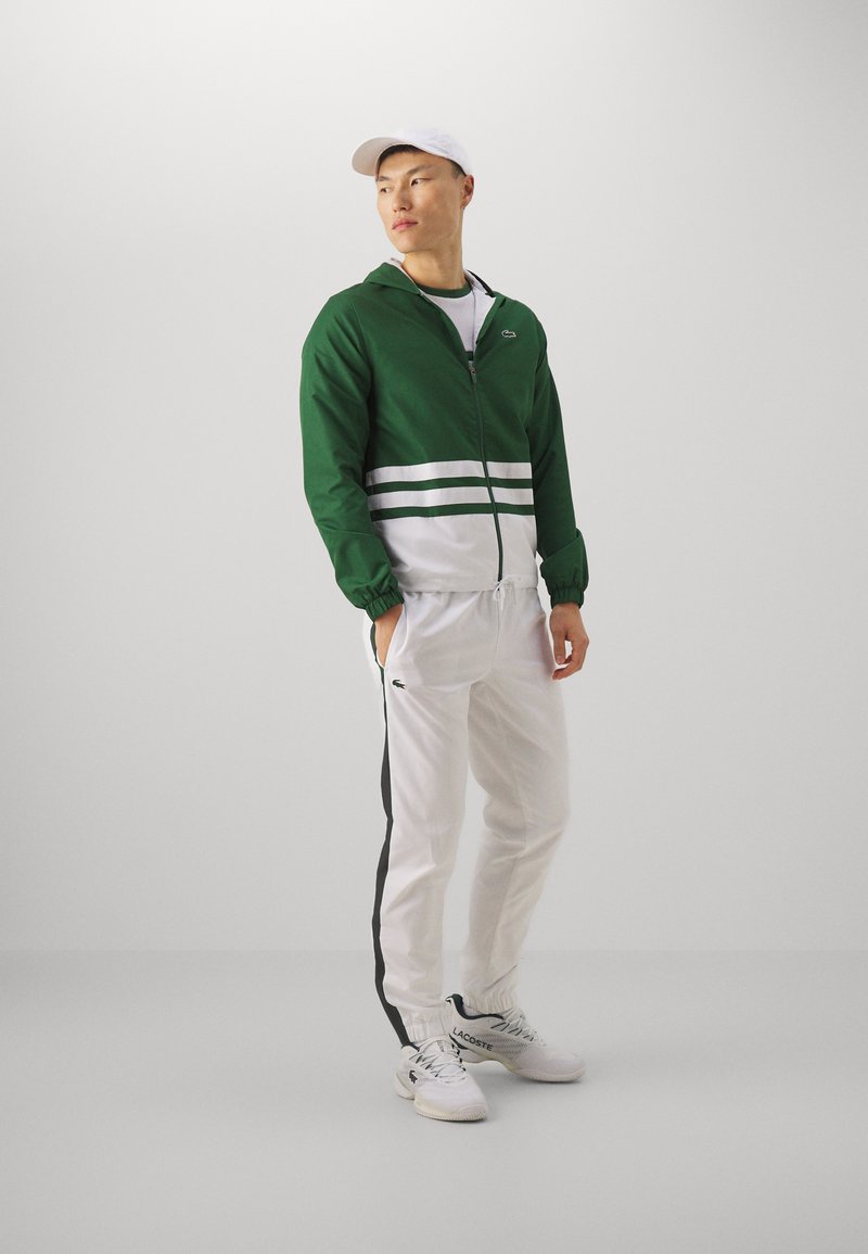 Chaqueta verde con cremallera y franjas horizontales blancas, camiseta blanca, pantalones blancos con franjas laterales negras y zapatillas blancas con detalles del logo.
