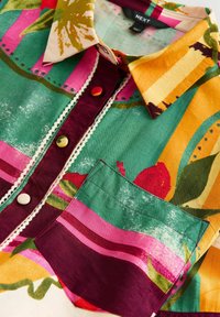 Chemise à col avec motifs floraux abstraits multicolores, poche avant, patte de boutonnage bordeaux avec bordure décorative et boutons recouverts de tissu.