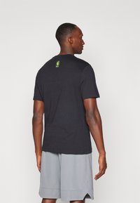 Camiseta de algodón negra con un logo verde de la NBA en la parte posterior. La camiseta tiene mangas cortas y un ajuste estándar, combinada con pantalones cortos grises.